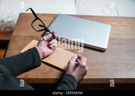 Hands writing in a notebook, holding glasses near a laptop on a wooden desk Stock Photo