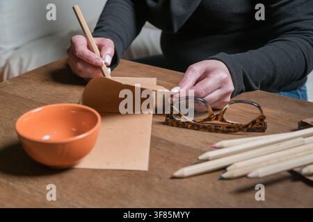 Close-up of hands folding a kraft paper envelope surrounded by craft tools on a wooden desk. Stock Photo
