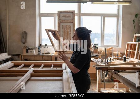 Female carpenter holds part of antique window frame in hands examining quality of wooden part. Stock Photo