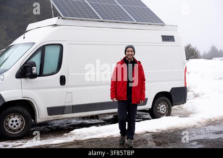 Young man smiling at the camera, standing in front of his white camper van with solar panels on the roof enjoying sustainable travel during winter Stock Photo