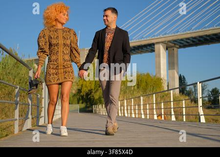 Diverse couple walking and holding hands on a bridge, enjoying outdoor leisure time Stock Photo
