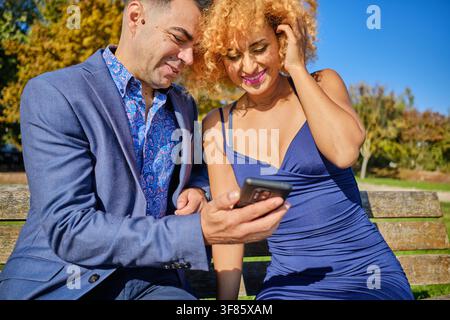 Young couple enjoying digital content while laughing together on a park bench outdoors Stock Photo