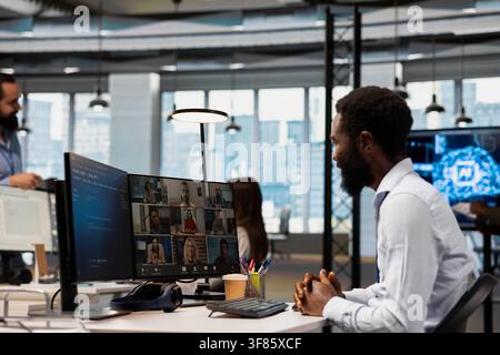 Smiling programmer in tech company office talking with team in videocall, doing brainstorming to protect against data loss. Jolly remote workers doing AI coding during internet videoconference Stock Photo