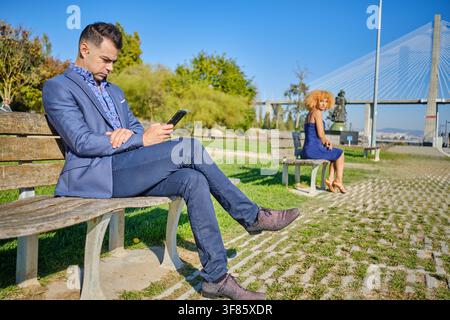 Man focused on phone, sitting on park bench, ignoring woman sitting on another bench Stock Photo