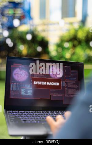 Close up of laptop displaying red system hacked alert over security windows, illustrating cybersecurity breach, malware warning and compromised digital system outdoors. Stock Photo