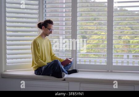 Man Reading Book While Sitting on Window Seat at Home Stock Photo