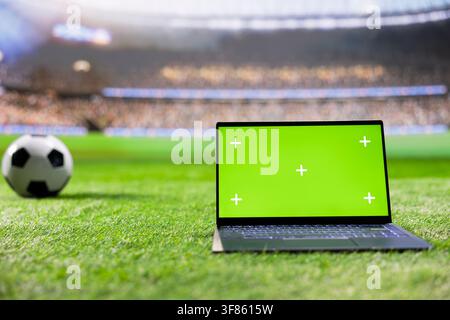 Chroma key isolated laptop screen placed at low angle on grass with soccer ball nearby and crowd behind in background. Copy space for screen replacement or editable screen. Stock Photo
