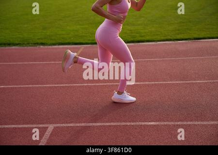 Close up of female runner legs mid-air on red stadium track. Athletic woman in pink leggings and white sneakers sprinting outdoors. Sports, dynamic ca Stock Photo