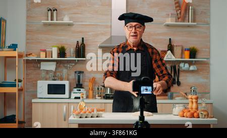 Inside the kitchen work area, the senior man records a baking demonstration for online viewers as camera equipment stands beside the kitchen worktop. Stock Photo