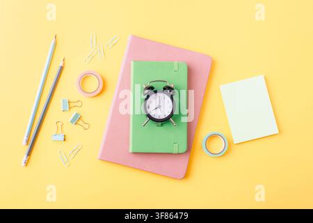 Green notebook and alarm clock surrounded by colorful stationery on a yellow background in a creative flat lay. Back to school concept. Top view, flat Stock Photo