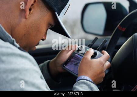 Young man in a cap sits in a car working the controls of a drone, the screen glowing in his hands. Stock Photo