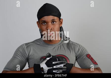 Studio portrait of a young Black man in a durag and grey T-shirt wearing padded gloves, arms folded on a grey backdrop. Stock Photo