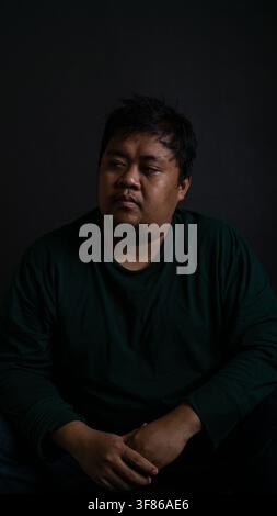A thoughtful portrait of a Southeast Asian male looking away while sitting against a solid dark black background, The adult Indonesian individual wear Stock Photo