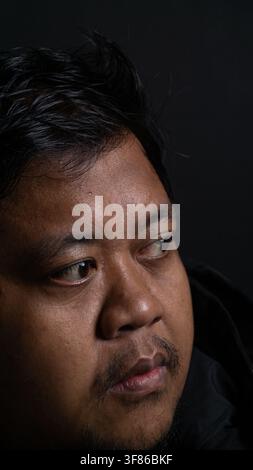 A dramatic side profile portrait of an adult Southeast Asian male looking away to the left side with a highly focused expression on a dark black backg Stock Photo