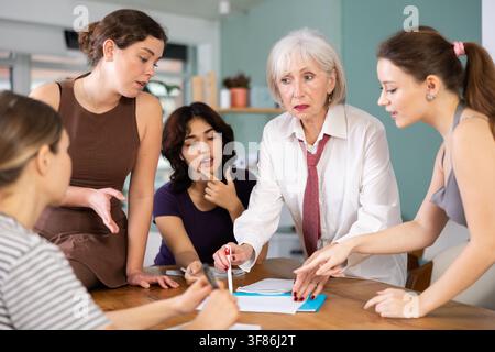 Emotional female employees and colleagues of business company discussing problems and plans while sitting at table in office Stock Photo