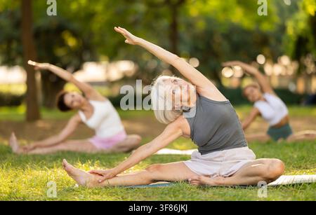 Active mature woman with group doing yoga position while sitting on mat at city park together Stock Photo