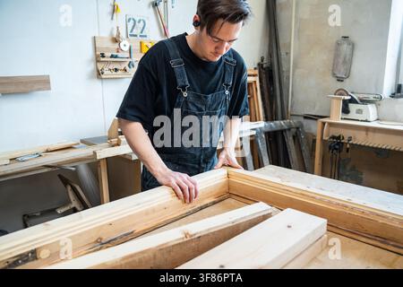 Craftsman connecting parts of refurbished antique wooden window frame at workbench in workshop. Stock Photo