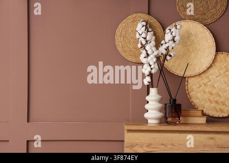 Modern home decor with cotton branches in a white vase and reed diffuser on a wooden cabinet against a brown wall, Stacked books add to the relaxed an Stock Photo