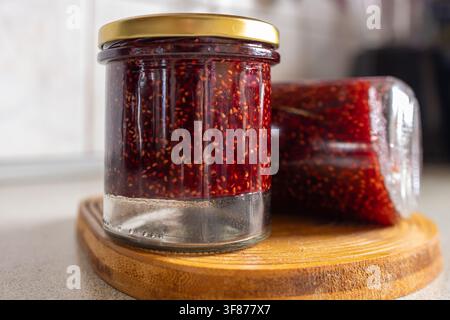 Close up details of transparent glass jars filled with bright red wild raspberry jam sitting on a round wooden surface in a domestic kitchen Stock Photo