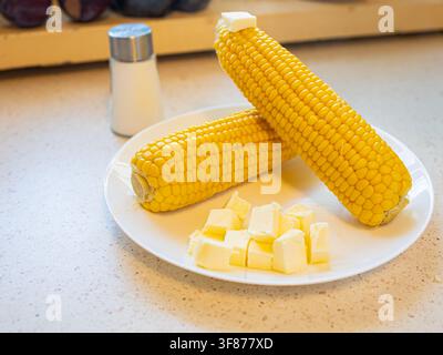 Two boiled sweet corn cobs with melting butter cubes on a yellow plate with a vintage glass salt shaker standing blurred in the background Stock Photo