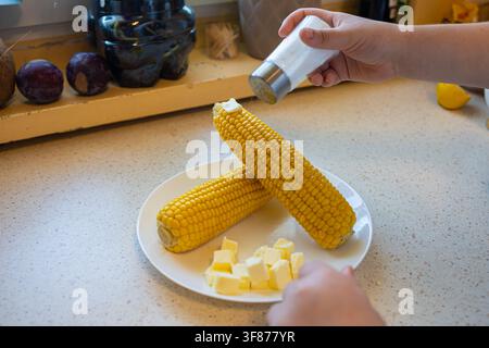 Close up of a hand shaking salt from a shaker onto two boiled sweet corn cobs served with melting butter cubes on a yellow plate in a kitchen Stock Photo