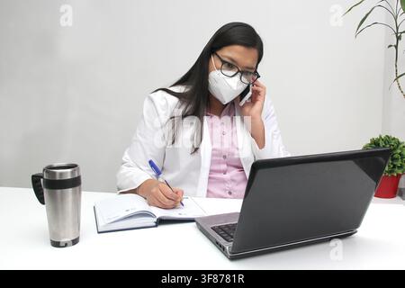 Latina doctor woman works in her office very busy uses her cell phone to answer messages, calls and video calls from patients Stock Photo