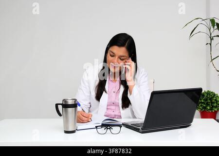 Latina doctor woman works in her office very busy uses her cell phone to answer messages, calls and video calls from patients Stock Photo