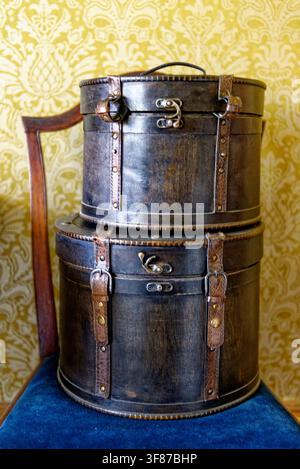 A pair of stacked, weathered dark leather hat boxes featuring vintage brass latches and straps, resting on a blue velvet chair at Lanhydrock House, Co Stock Photo