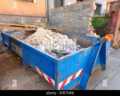 Large blue industrial waste container filled with construction debris and rubble from a demolished brick wall next to a house Stock Photo