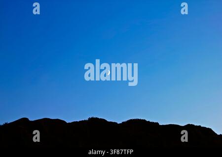 A waxing moon in a clear sky over silhouetted mountains. Stock Photo