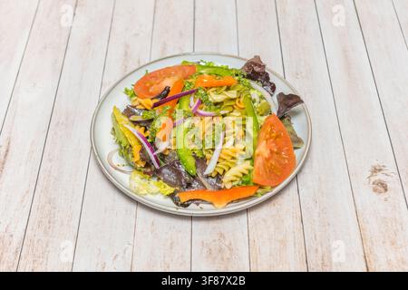 Vibrant fusilli pasta salad with mixed greens, tomatoes, bell peppers, red onion, and pesto served on a white plate. Stock Photo