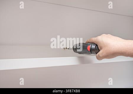 Hand using cordless power drill to install screw into white cabinet frame during furniture assembly. Stock Photo