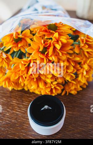 A sparkling diamond engagement ring in an open white box sits in front of a vibrant bouquet of bright orange chrysanthemums on a wooden table. Stock Photo