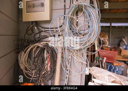 Russia, Saint Petersburg, 23.08.2025 Bundle of electrical cables and wiring hanging in mess. Disorganized cabling in garage workshop or utility room. Stock Photo