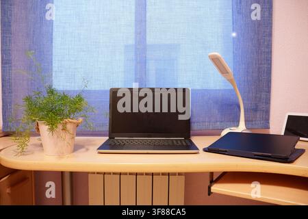 Minimal home office workstation featuring closed laptop on wooden desk beneath sheer curtained window, tablet, stylus, adjustable lamp, potted plant Stock Photo