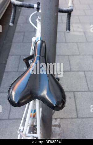 A bicycle seat is locked to a metal pole on a sidewalk in a busy city. The sun is shining, and people are walking by. The street has bricks and a bicy Stock Photo