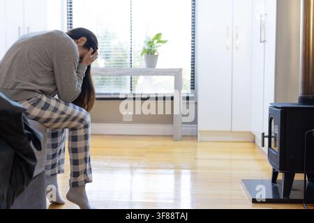 Woman sitting on couch wearing plaid pajama pants holding face in hands at home, copy space. Solitary, contemplative, cozy, loungewear, woodstove, bli Stock Photo