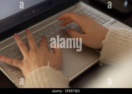 Woman typing on silver laptop on dark wood desk wearing fuzzy sweater, showing pink nails. Hands, keyboard, closeup, warm, cozy, wood, focus Stock Photo