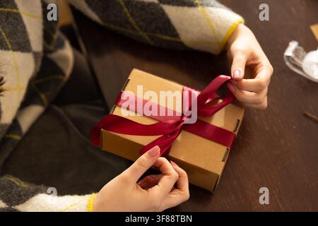 Adult female in argyle sweater tying burgundy ribbon on box at wooden table with tape dispenser. Gift, present, bow, wrap, satin, knit, cozy Stock Photo