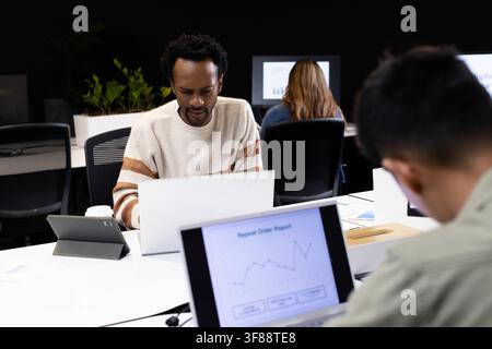 Diverse coworkers working on laptops and tablets at open-plan office with man in cream sweater. Workspace, coworking, collaboration, focus, business, Stock Photo