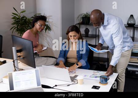 Office team collaborating around white desk at modern office reviewing laptop and printed charts. Collaboration, colleagues, analytics, graphs, report Stock Photo