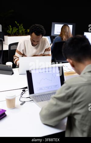 Diverse coworkers analyzing laptop charts at office desk with tablet, man wearing cream sweater. Data, analytics, reports, graphs, monitors, workspace Stock Photo