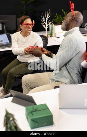 African American coworkers exchanging small red gift wearing reindeer glasses in office with laptop. Collaboration, colleagues, celebration, festive, Stock Photo