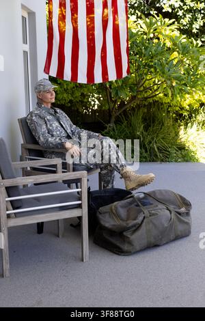 Midlife woman sitting on porch chair wearing camo uniform tan boots, olive duffel, black pack, flag. Patio, outdoor, greenery, garden, sunlight, seren Stock Photo