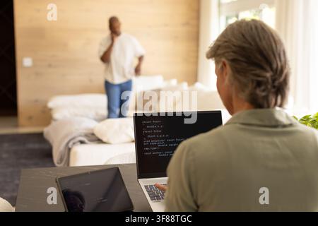 Couple sharing living room, woman coding on laptop at table, man talking on smartphone, copy space. Remote, programming, computer, home, workspace, da Stock Photo