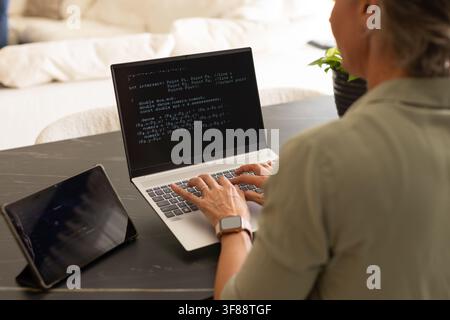Woman coding on laptop in home workspace wearing olive shirt tablet smartwatch plant, copy space. Programmer, developer, homeoffice, livingroom, minim Stock Photo