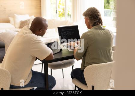 Diverse couple working on laptop with terminal and tablet at small black table in bedroom. Collaboration, remote, coding, programming, homeoffice, min Stock Photo