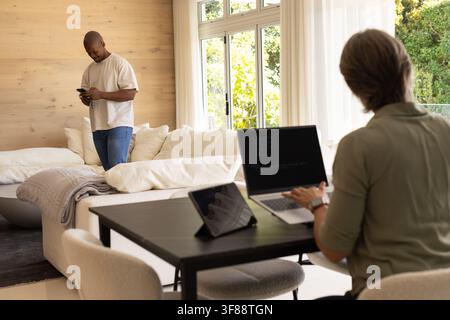 Diverse couple working in living room, woman coding on laptop with tablet, man checking smartphone. Computer, code, remotework, homeoffice, workspace, Stock Photo