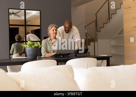 Diverse couple leaning over laptop on stand at dining table, senior woman with tablet and plant. Homeoffice, pair, collaboration, contemporary, minima Stock Photo