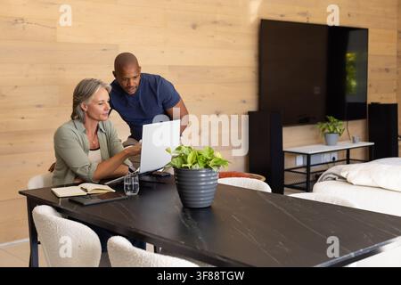 Diverse couple working together at home using laptop on black dining table with notebook, plant. Collaboration, remote, workstation, minimalist, moder Stock Photo